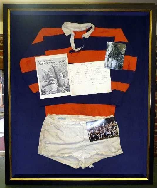 A framed display of a blue and orange rugby jersey, white shorts, a team photo, and a commemorative pamphlet on blue felt.