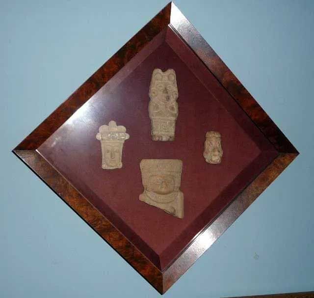 Four small, carved stone-like artifacts mounted on a dark red fabric background inside a diamond-shaped wooden frame.