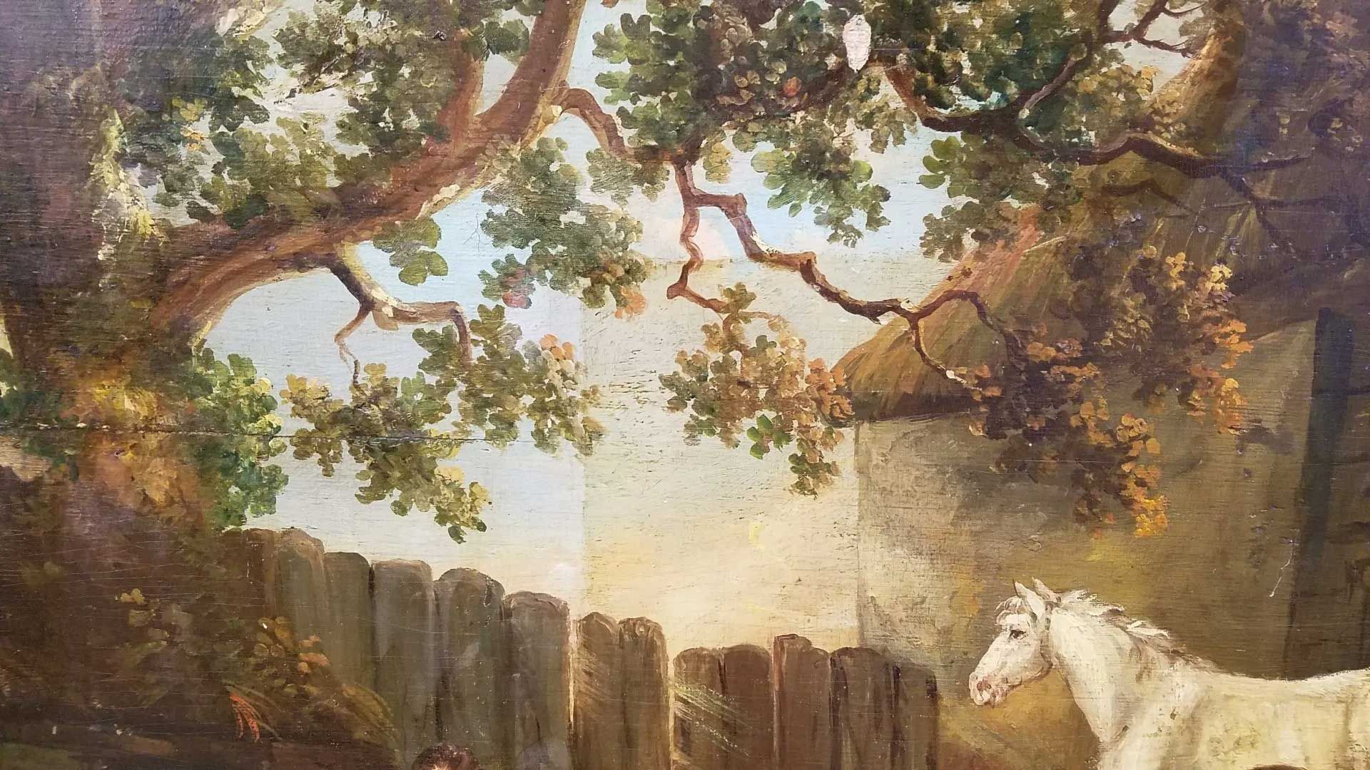 A white horse stands by a wooden fence near a stone building under the canopy of a large, leafy tree.