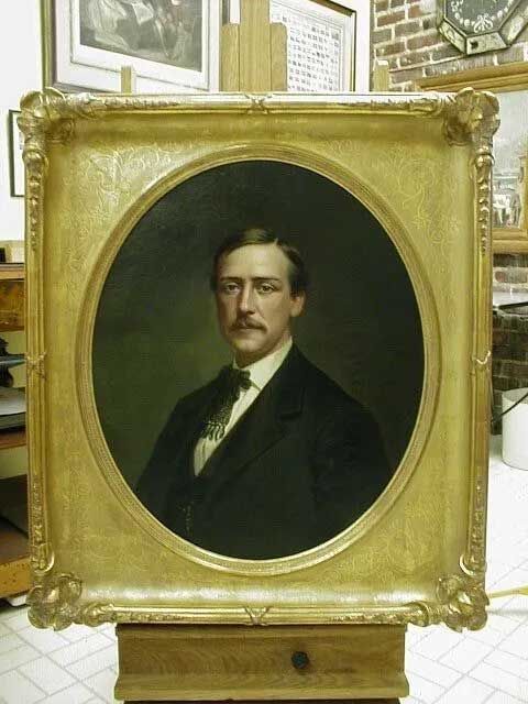 A portrait of a person with a mustache in a dark suit, displayed in an ornate gold oval frame on a wooden easel.