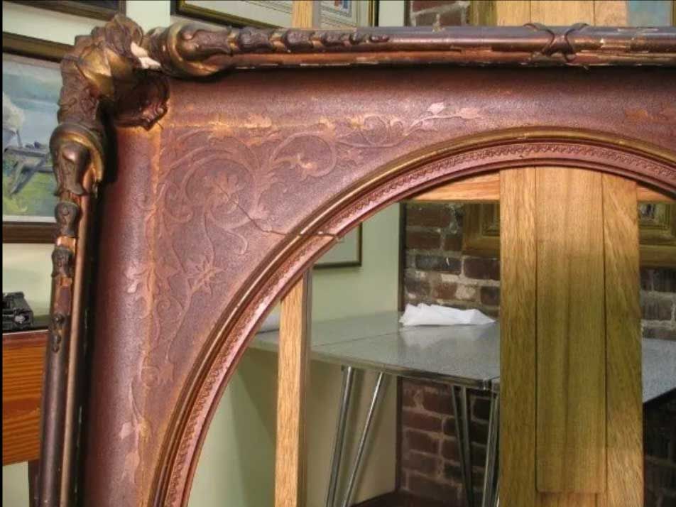 A close-up of a weathered, dark red ornate wooden picture frame with gilded gold detailing, mounted on a wooden easel.