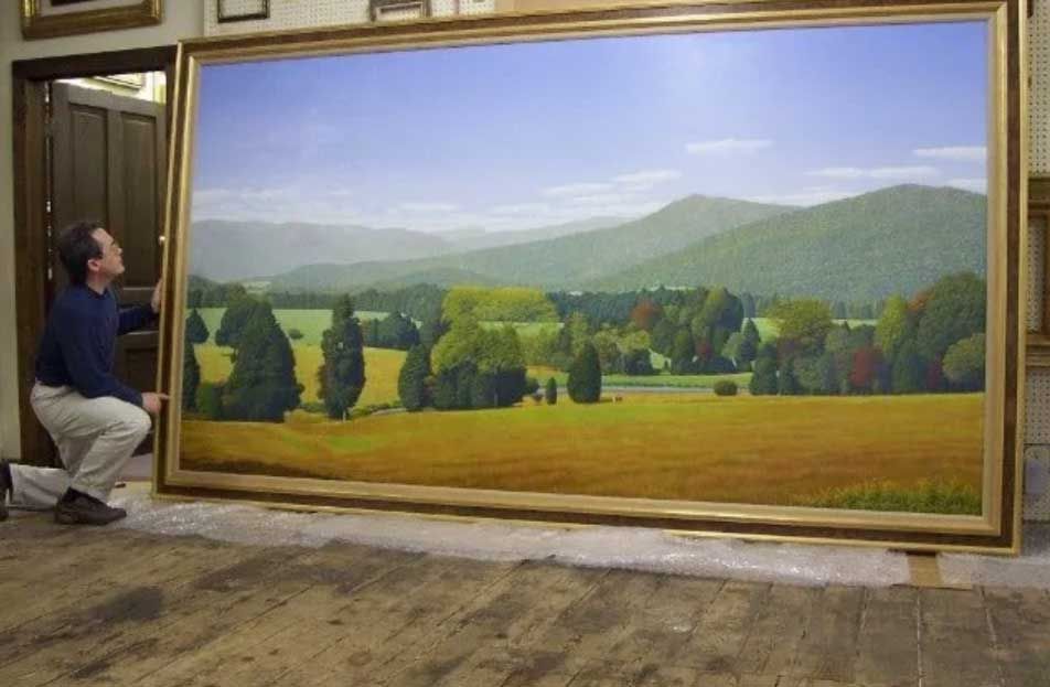 A person kneels beside a large, framed landscape painting of rolling green hills, forests, and fields under a blue sky.