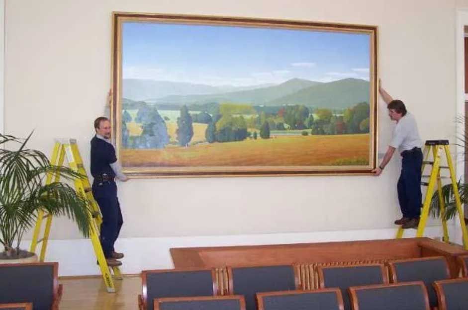 Two workers on step ladders hang a large landscape painting on a beige wall above seating in a room.