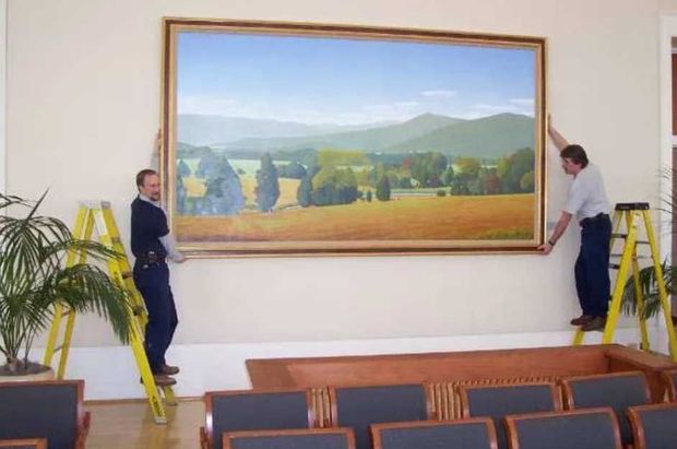 Two individuals standing on yellow step ladders hang a large, framed landscape painting on a beige wall in a room.