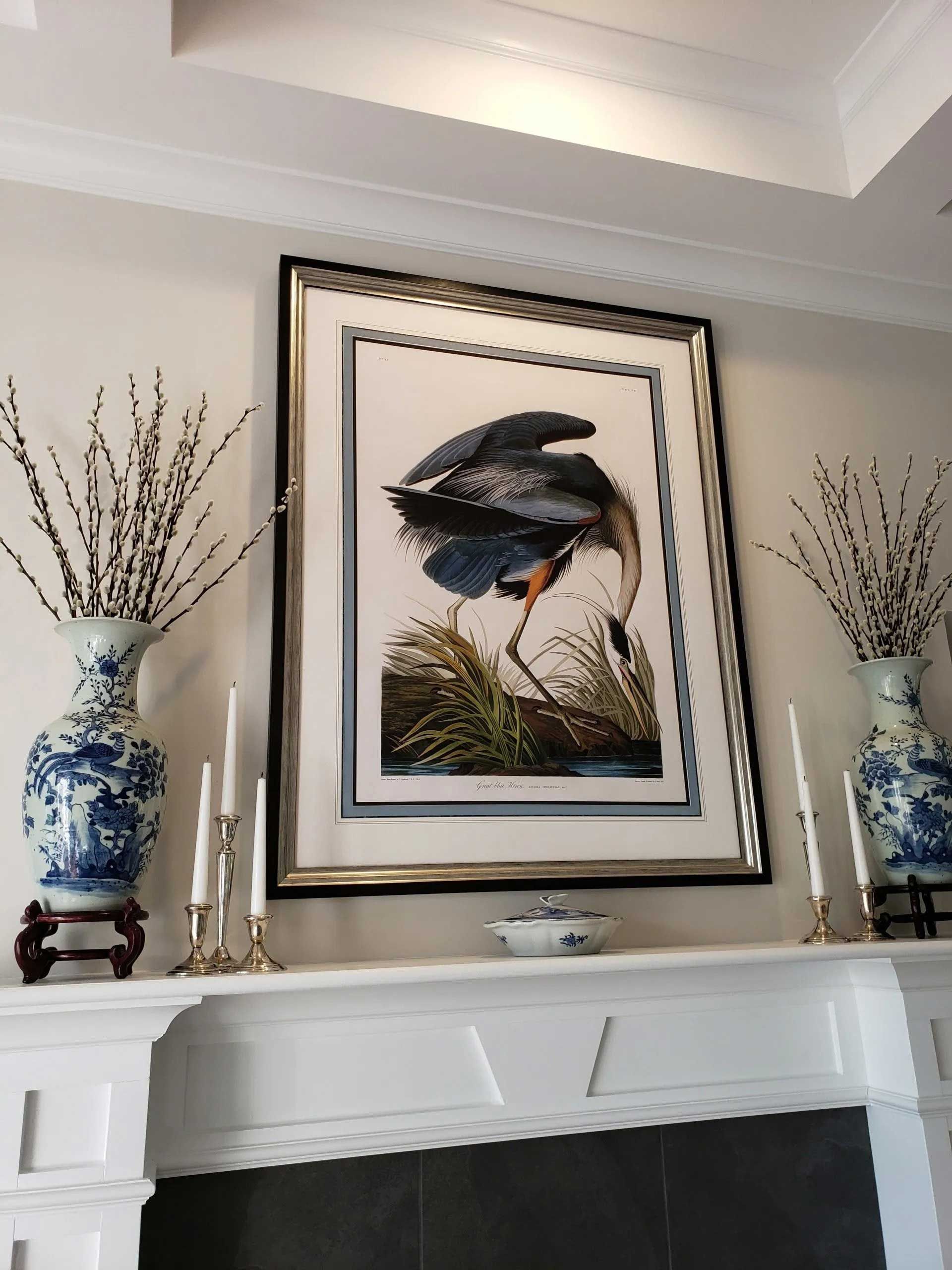 A framed print of a blue heron hangs above a white mantelpiece, flanked by two blue and white vases with bare branches.