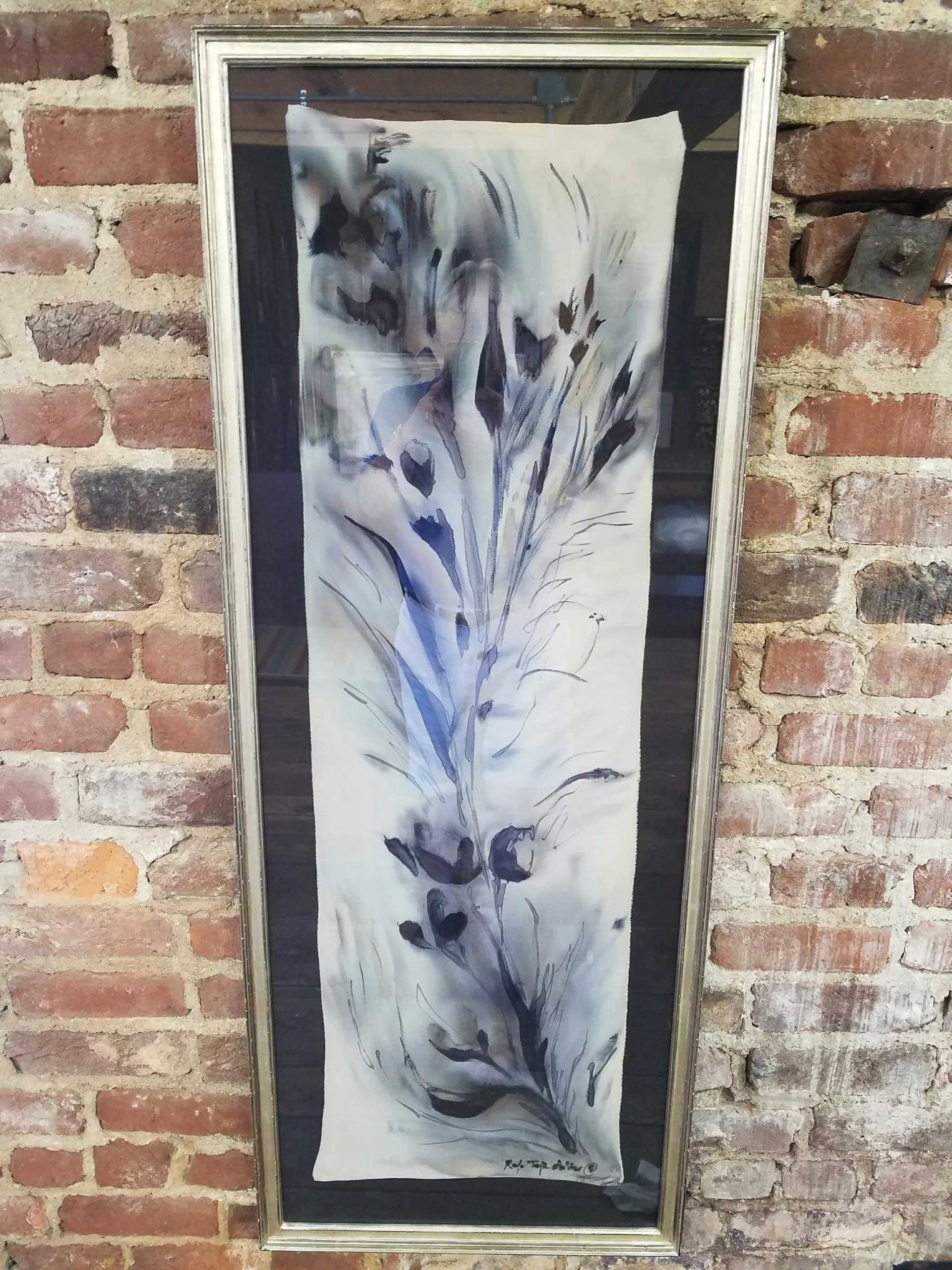 A long, framed vertical painting of abstract dark blue and grey feathery shapes on a cream background, on a brick wall.