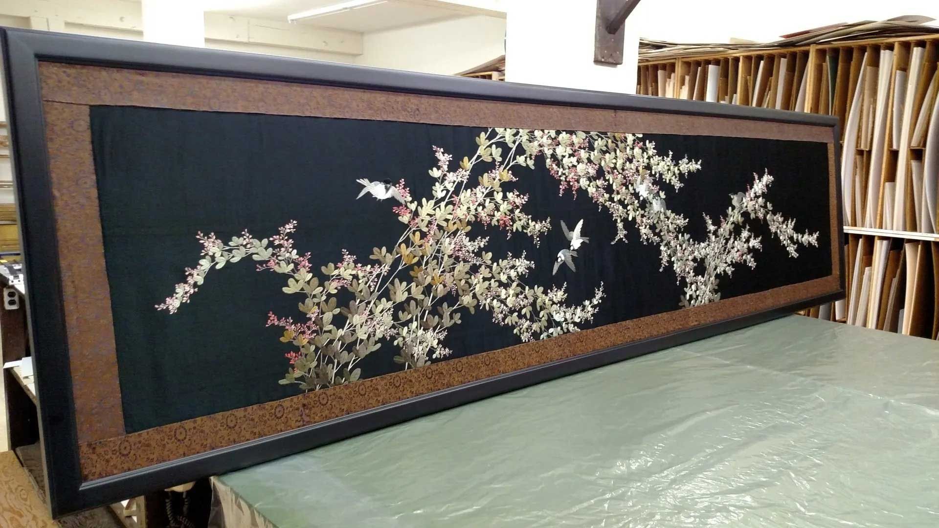 A framed, wide horizontal embroidery on black fabric featuring white and pink cherry blossoms with birds, in a workshop.