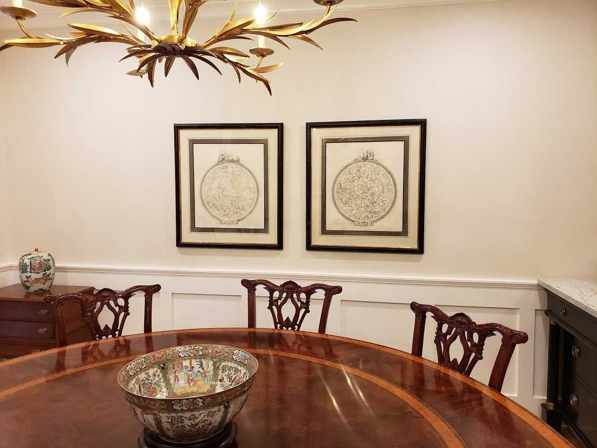 A dining room featuring a round wooden table, chairs, a decorative bowl, a brass chandelier, and two framed wall prints.