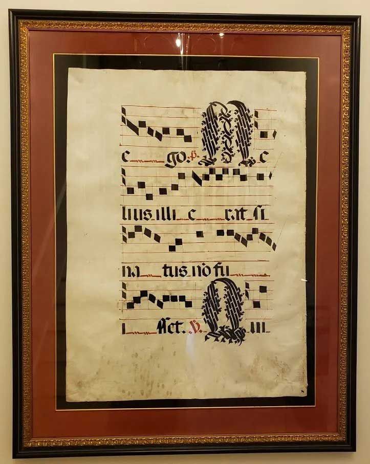 A framed piece of medieval musical notation on parchment with black square notes, red staff lines, and decorative letters.