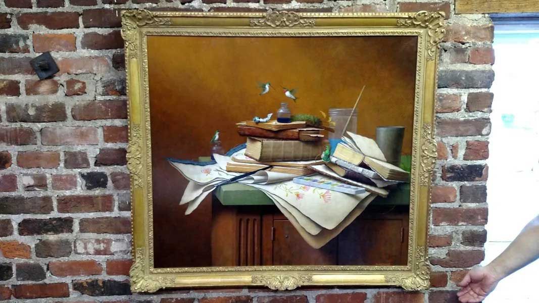 A still-life painting of books and papers on a wooden table, framed in gold, mounted on a rustic exposed brick wall.