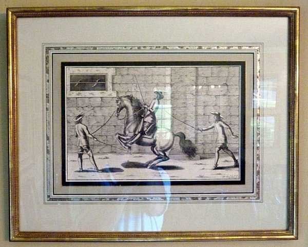 Framed black and white etching of a person on a rearing horse, being guided by two individuals in a walled courtyard.