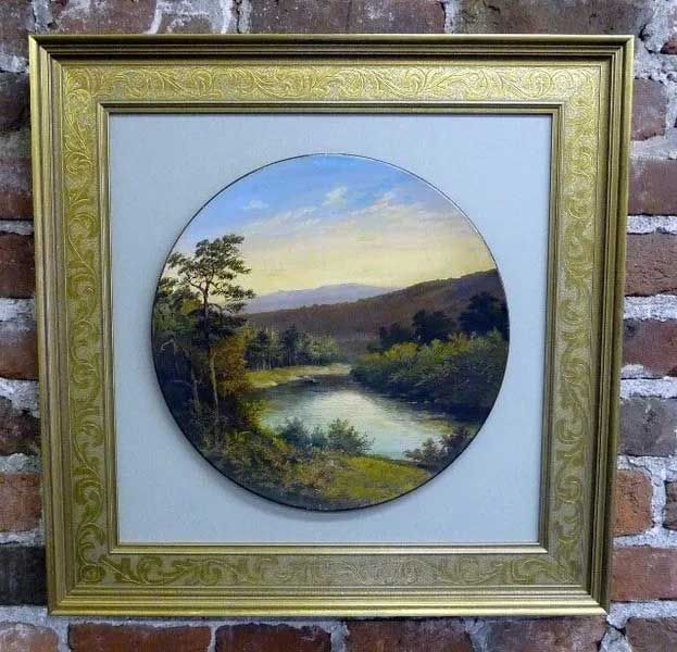 A circular landscape painting of a lake surrounded by trees and hills, displayed in a square, ornate gold frame.