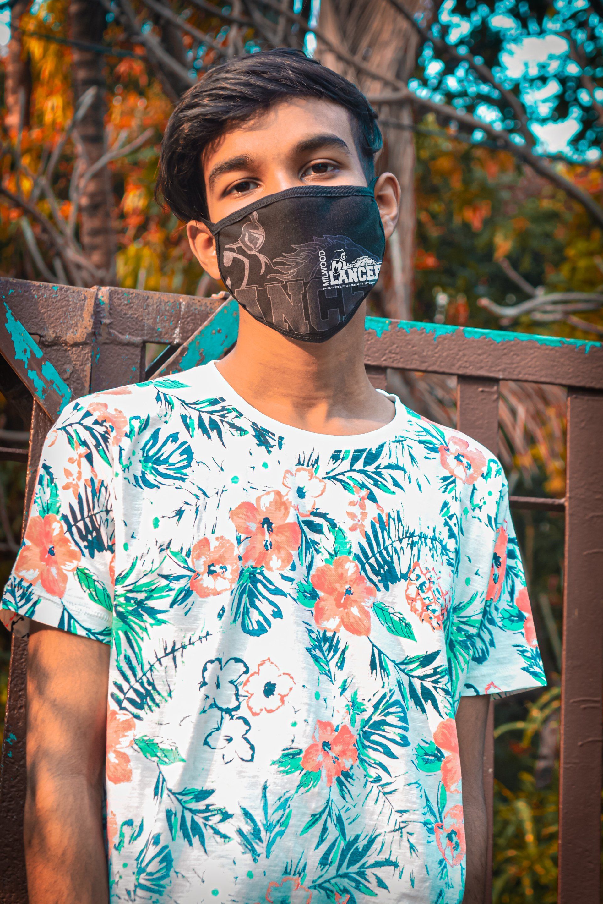 A young man wearing a face mask and a floral shirt is standing in front of a wooden fence.