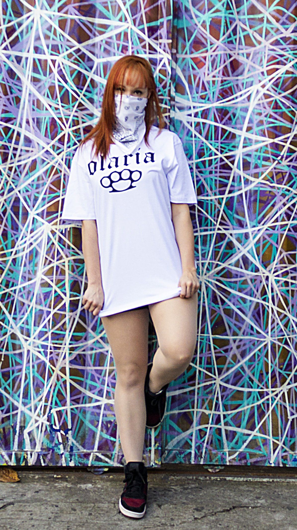 A woman wearing a mask and a white t-shirt is standing in front of a graffiti wall.