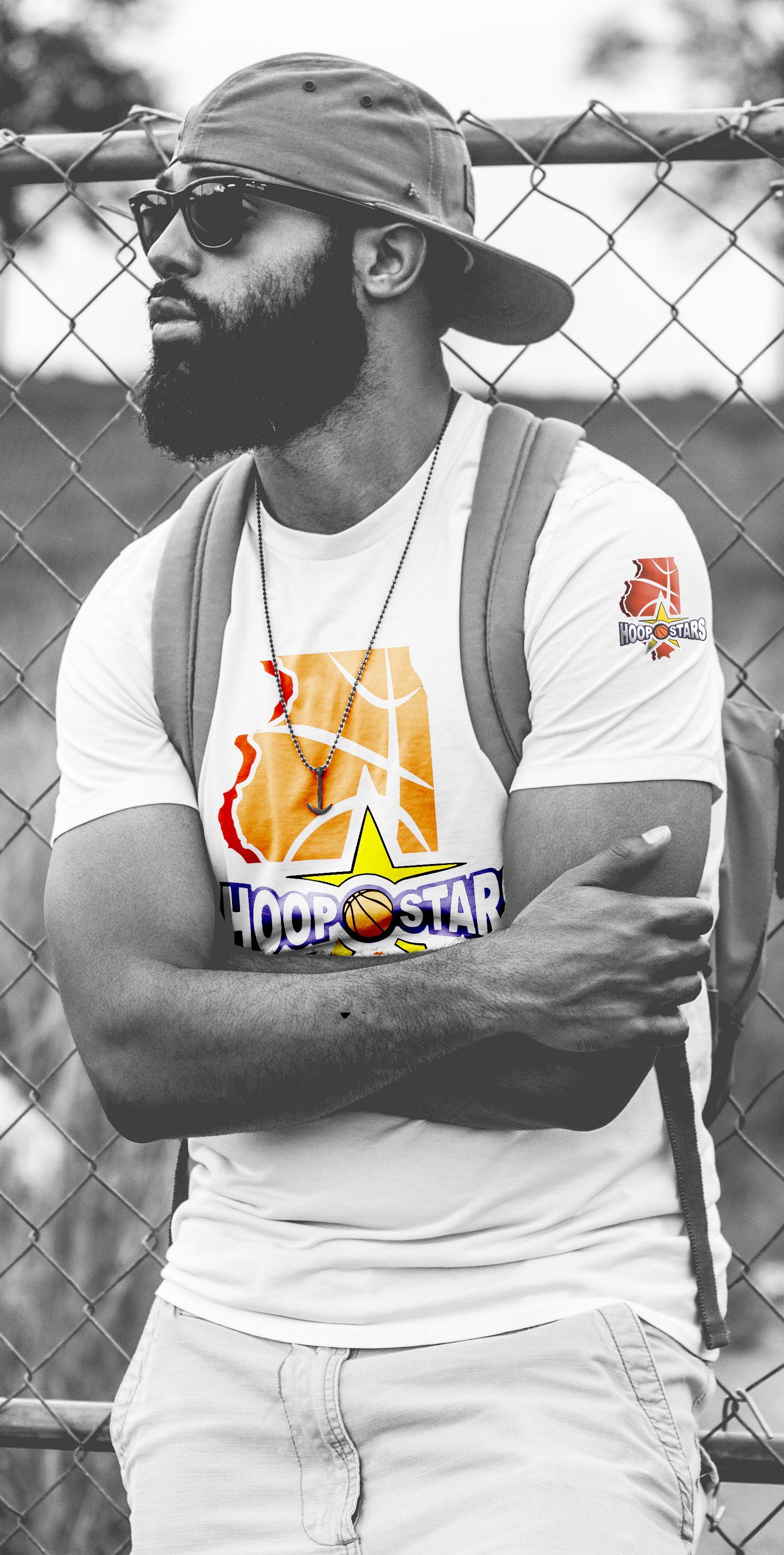 A man with a beard wearing a hat and sunglasses is standing in front of a chain link fence.