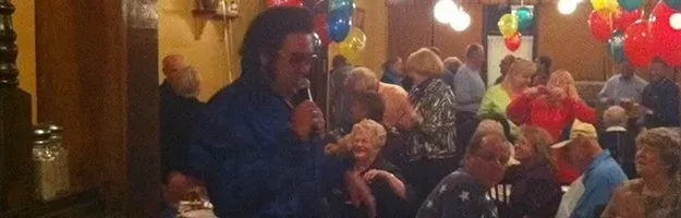Elvis impersonator performing for a crowd at a party, with balloons in the background.