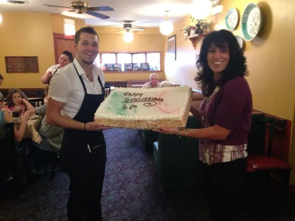 Server and woman holding a large rectangular birthday cake in a restaurant, smiles.