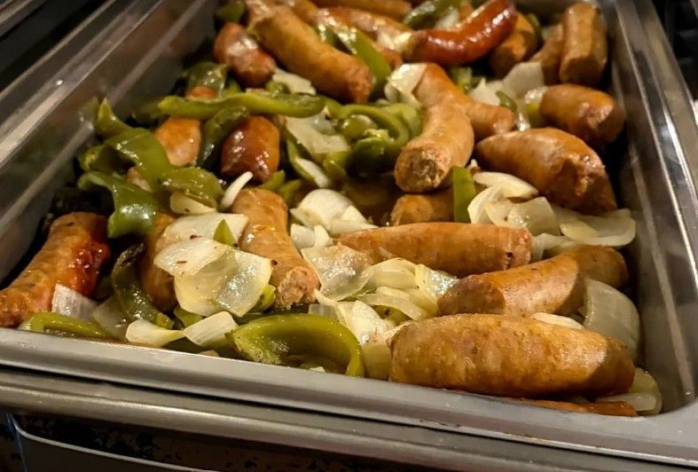 A metal serving tray filled with cooked sausages sautéed with sliced green bell peppers and onions.