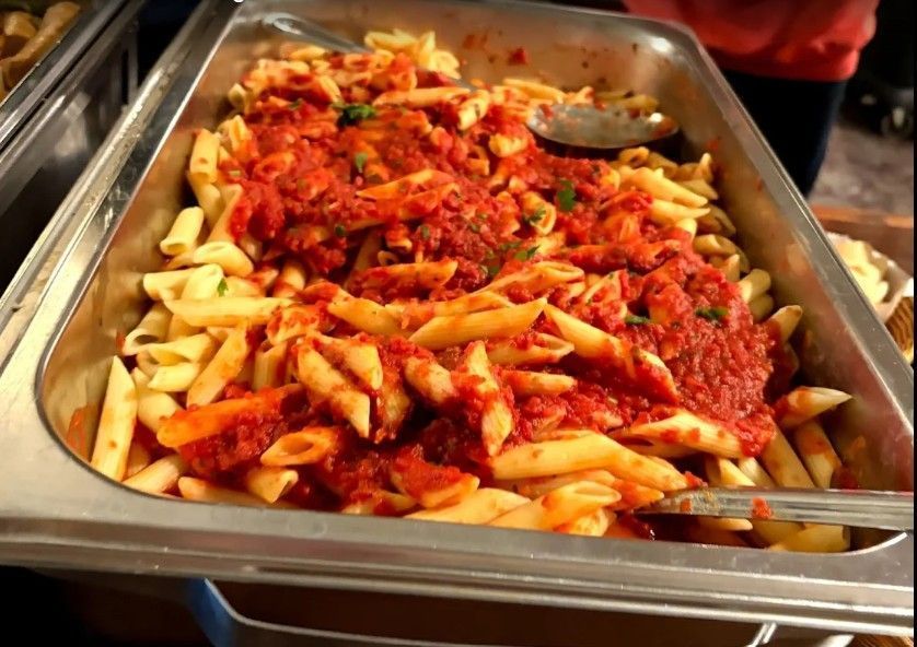 A metal catering tray filled with cooked penne pasta topped with a thick red tomato sauce and a serving spoon.