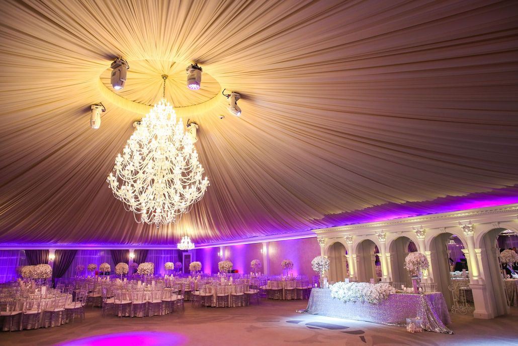 Elegant ballroom decorated for a wedding with a large chandelier and purple lighting. Tables are set.