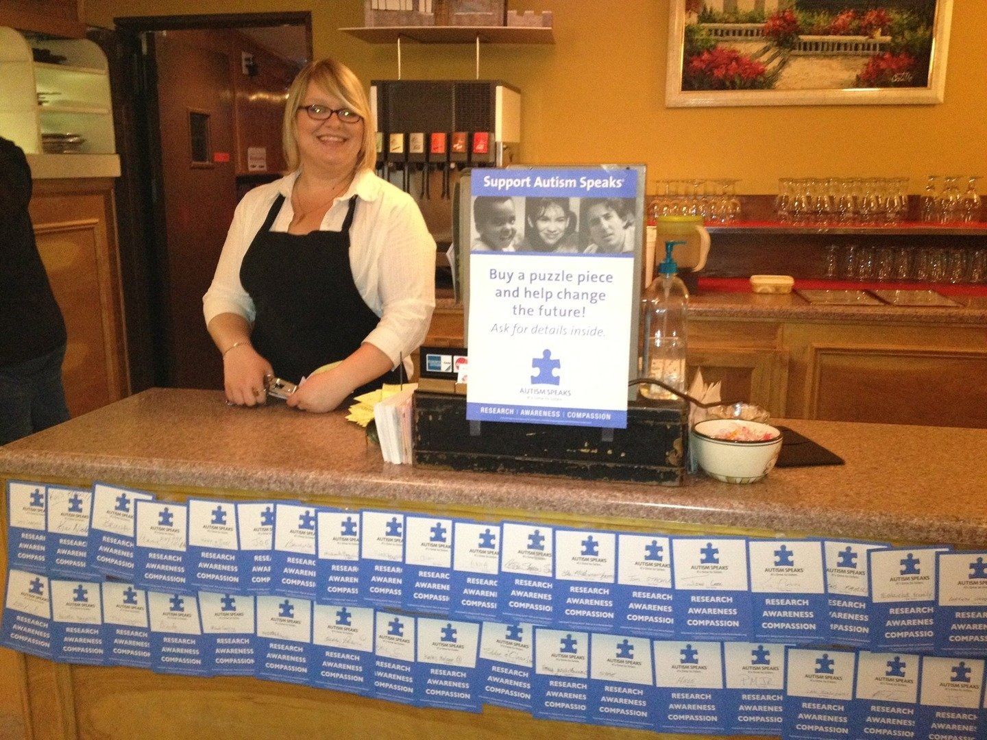 A smiling staff member stands behind a counter covered in Autism Speaks puzzle piece donation slips for a fundraiser.