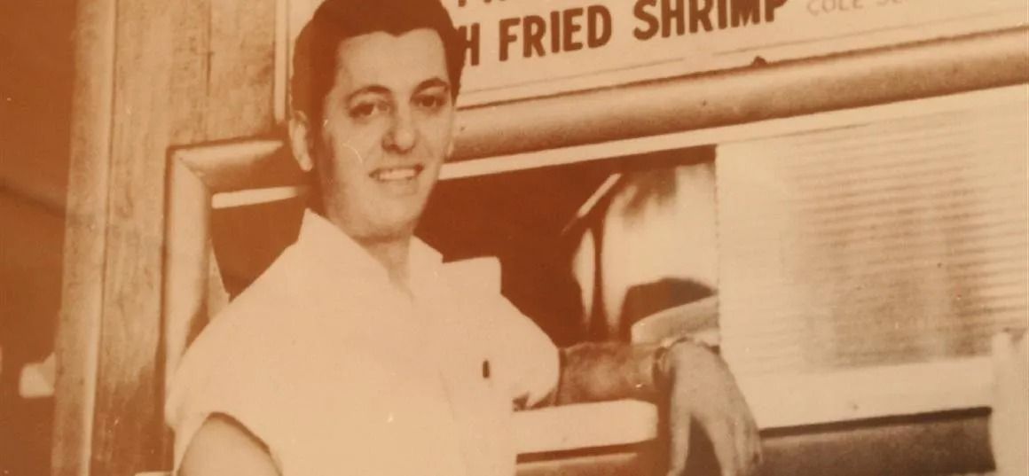 A smiling person in a light-colored shirt standing behind a counter with a sign that reads, 