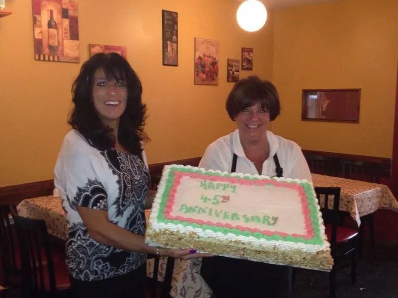 Two women holding a large cake celebrating a 45th anniversary, in a restaurant.