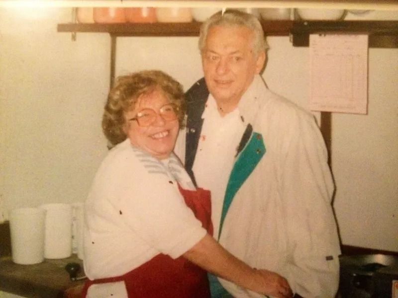 A smiling woman in a red apron embraces a man in a light jacket inside what appears to be a kitchen or pantry.