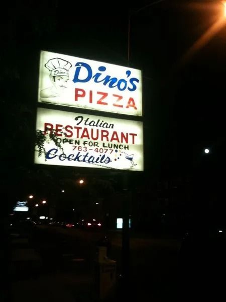 A glowing outdoor sign for Dino's Pizza and Italian Restaurant at night, including a phone number and mention of cocktails.
