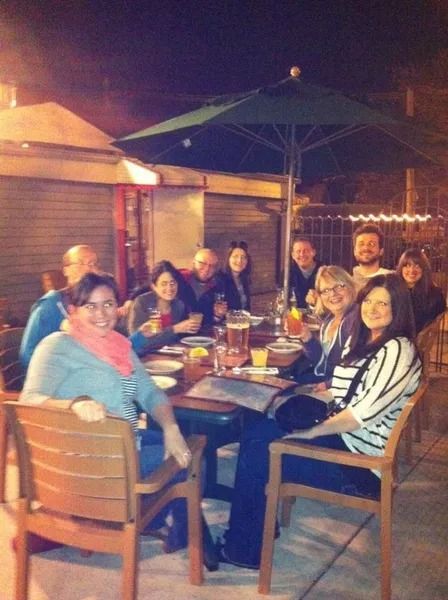 A group of friends sits around an outdoor patio table with drinks under an umbrella at night.