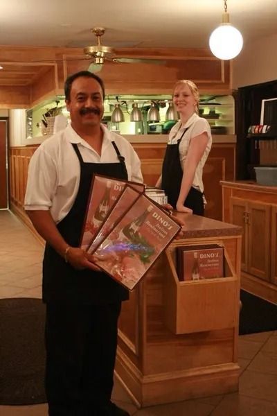 A restaurant employee holds menus in front of a counter while a smiling colleague stands in the background.