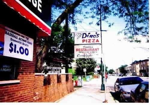 A street view of a brick building with a sign advertising $1 drinks, next to a sign for Dino's Pizza and Restaurant.