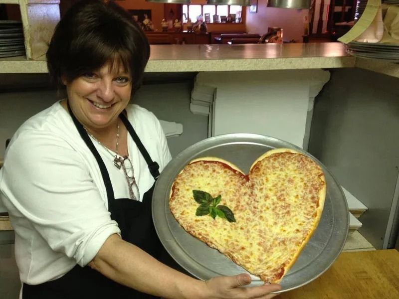Woman holding heart-shaped pizza on a tray in a restaurant. She smiles and has an apron on.
