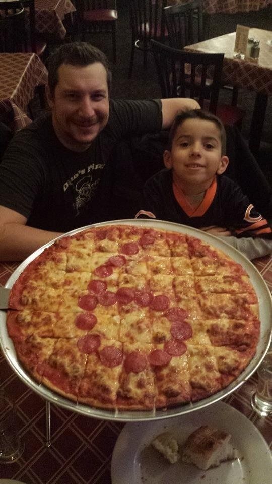 Man and child smiling with a large pepperoni pizza in a restaurant setting.