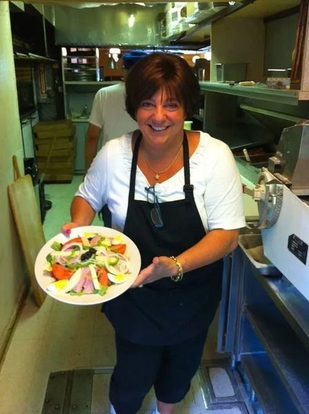 Woman in black apron holding a salad in a restaurant kitchen, smiling.