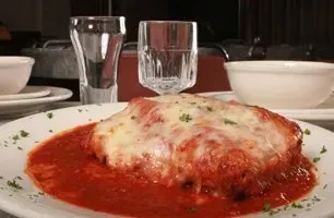 Chicken Parmesan on a white plate with red sauce, garnished with parsley, alongside two water glasses.