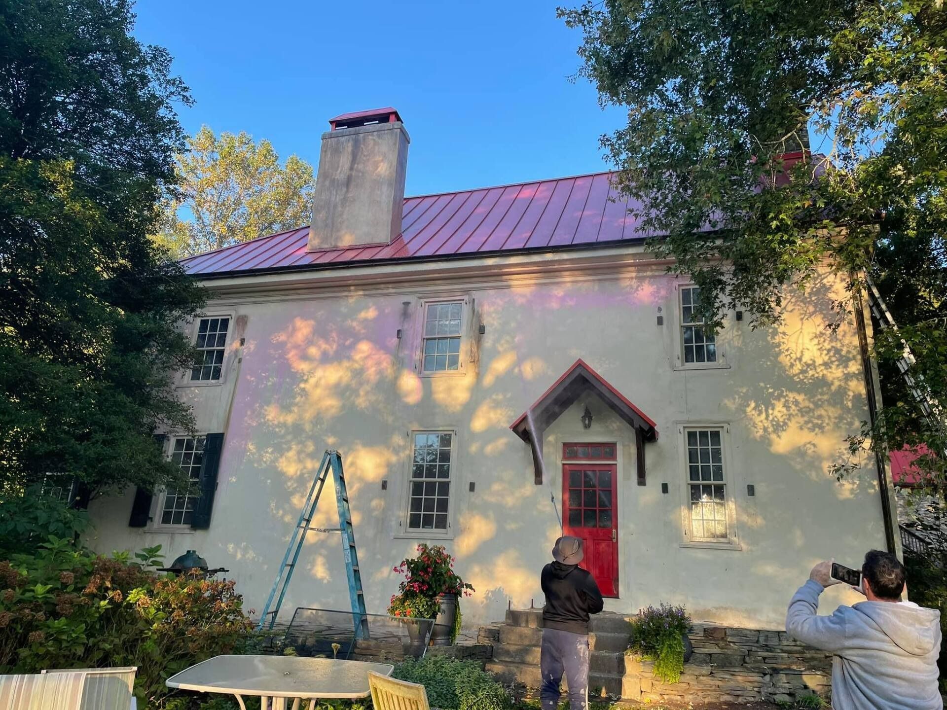 Two-story white house with red door and roof; people outside; a ladder leans against the building.