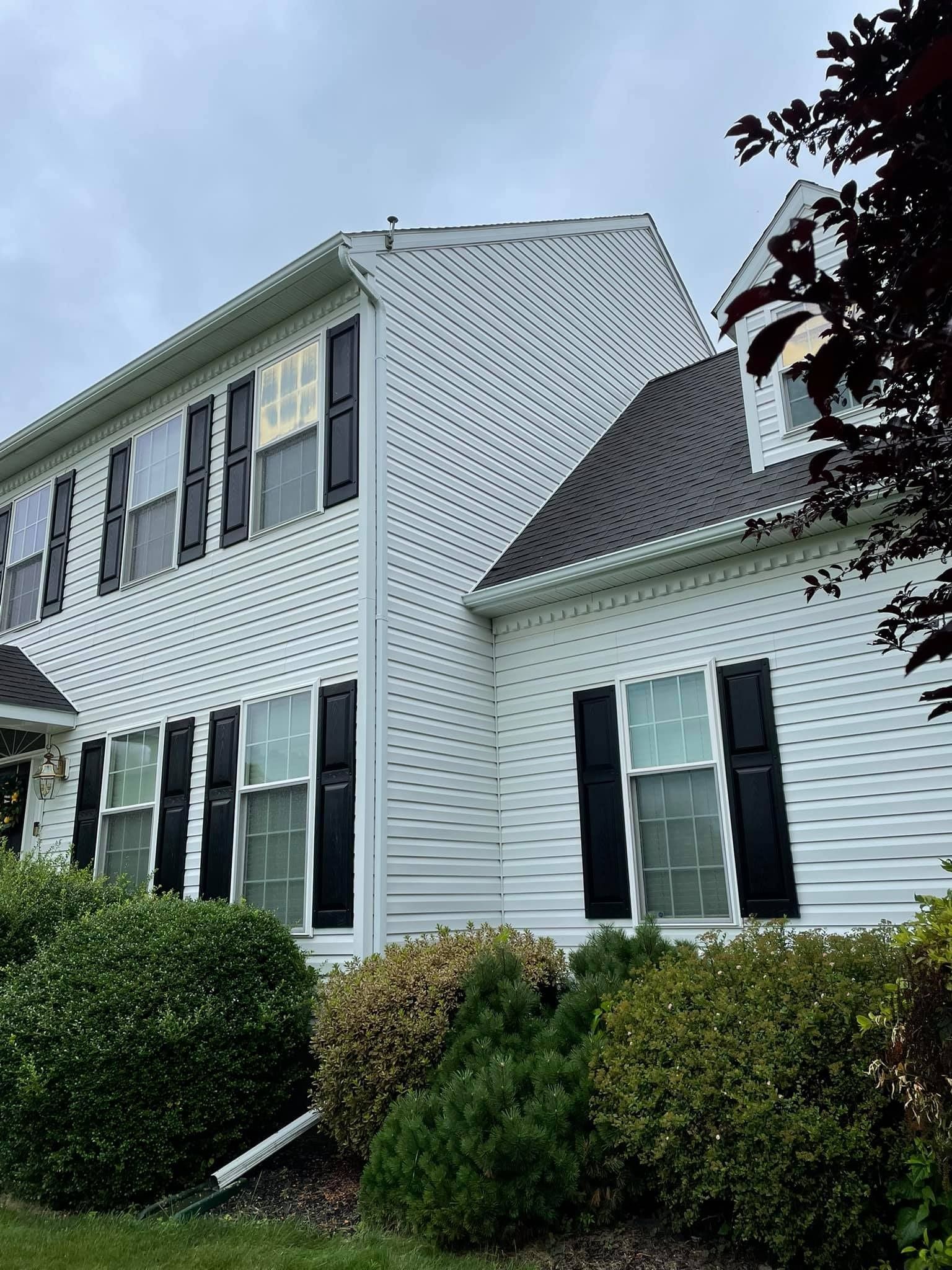 Two-story white house with black shutters, roof, and bushes. Overcast sky.