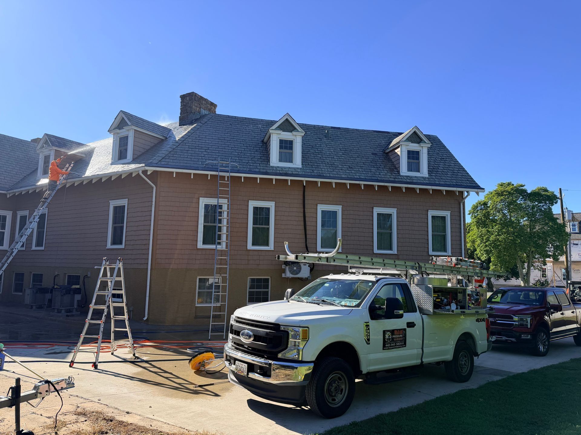 Brown building with multiple dormer windows, workers on ladders, a white pickup truck, and clear blue sky.