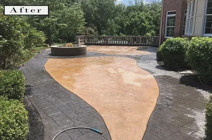 Backyard patio with brown concrete, black pavers, and a fire pit. Trees and a deck are in the background.