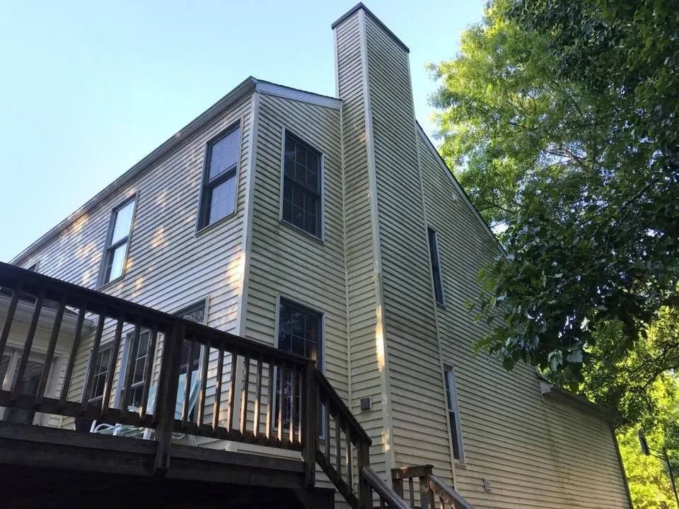 Two-story house with a wooden deck and a chimney, surrounded by green trees under a blue sky.