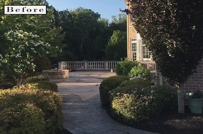 Pathway leading to a stone patio with a balustrade, surrounded by greenery and trees. Brick house visible.
