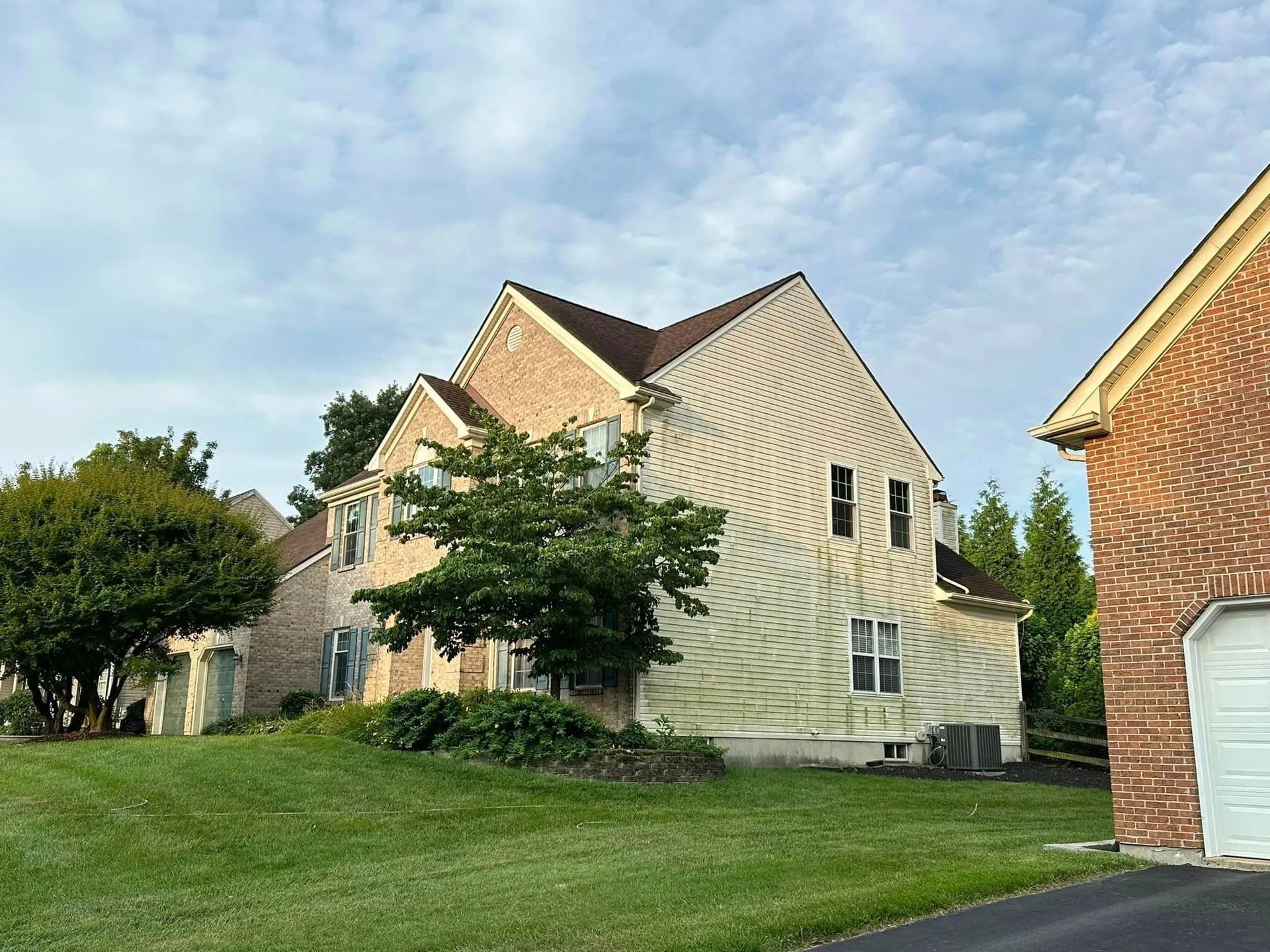 Two-story house with beige siding, brown roof, and brick facade, green lawn under a cloudy sky.