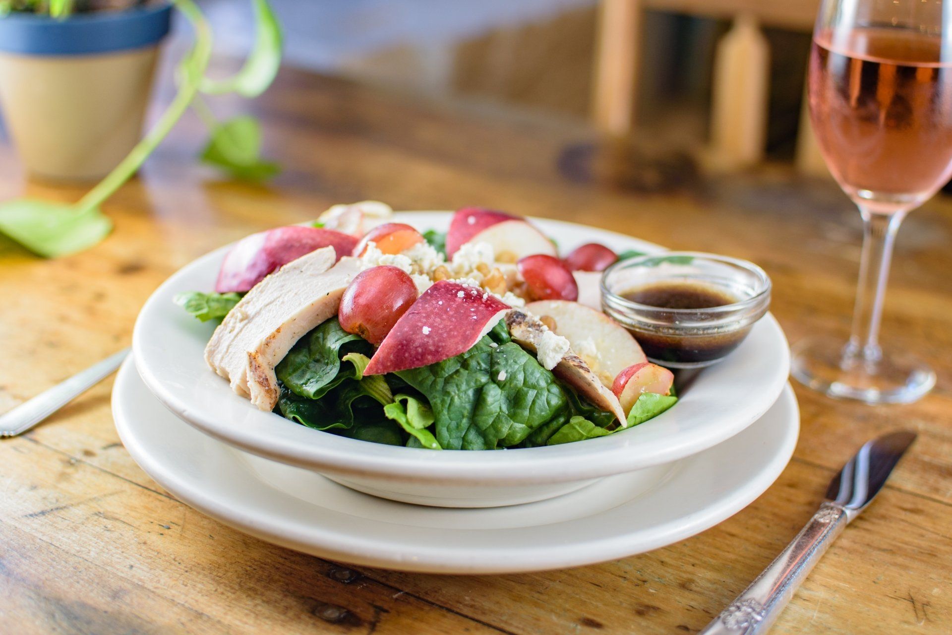 A bowl of salad with chicken , spinach , grapes and a glass of wine on a wooden table.