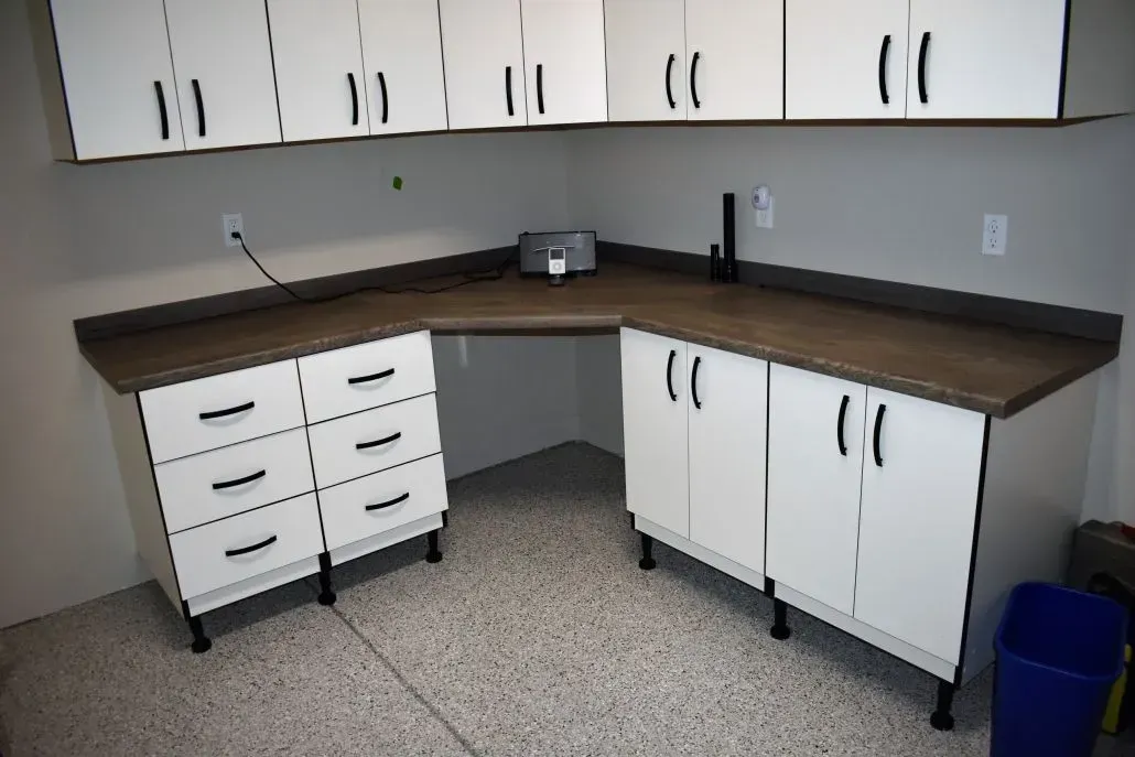 White cabinets with a dark countertop in a garage. A corner desk is below wall cabinets.