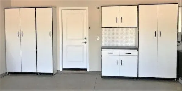 White storage cabinets in a garage. Door on the right, cabinets with black trim and doors. Gray floor.