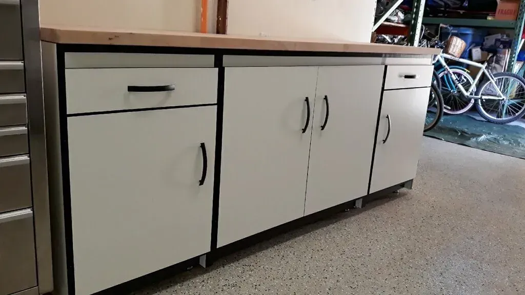 White cabinets with black handles and drawers, topped by a light wood counter, in a garage.
