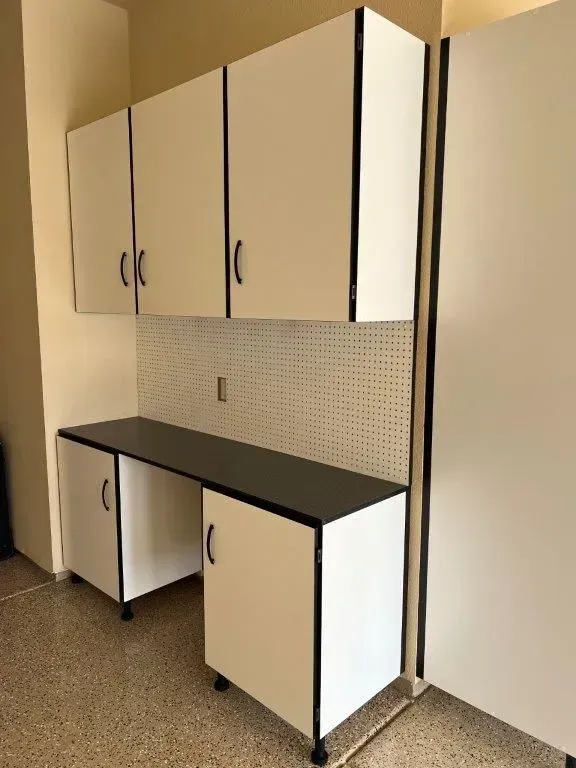 White and black storage cabinets in a garage. Includes overhead cabinets, a workspace, and a pegboard.