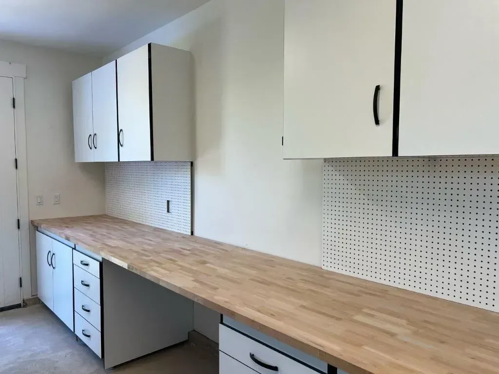 White cabinets and butcher block countertop in a garage. Pegboard backsplash, black hardware.
