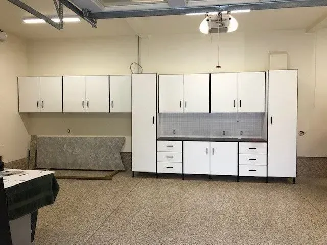White garage cabinets on a speckled floor. Cabinets include wall, tall, and base units. A large countertop leans against the wall.